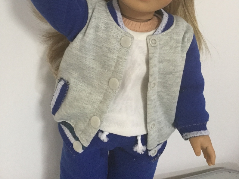 May include: A gray and blue doll outfit with a white shirt, gray jacket with blue sleeves, and blue pants. The jacket has a button closure and the pants have a drawstring.