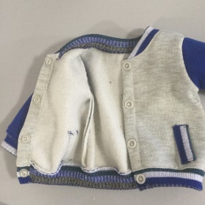 May include: A gray and blue varsity jacket with a zipper closure and snap buttons. The jacket has a white stripe on the sleeves and a pocket on the right side.