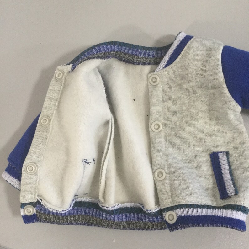 May include: A gray and blue varsity jacket with a zipper closure and snap buttons. The jacket has a white stripe on the sleeves and a pocket on the right side.
