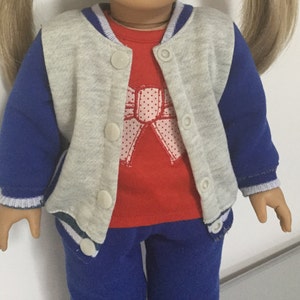 May include: A gray and blue doll outfit with a red shirt featuring a pink bow with white polka dots. The outfit includes a gray and blue baseball jacket, blue pants, and a red shirt.