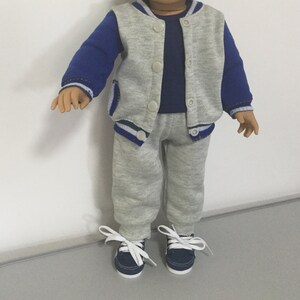 May include: A gray and blue tracksuit for a doll. The jacket has a blue and white stripe on the sleeves and a white stripe on the bottom. The pants are gray with a white stripe on the bottom. The doll is wearing navy blue sneakers.