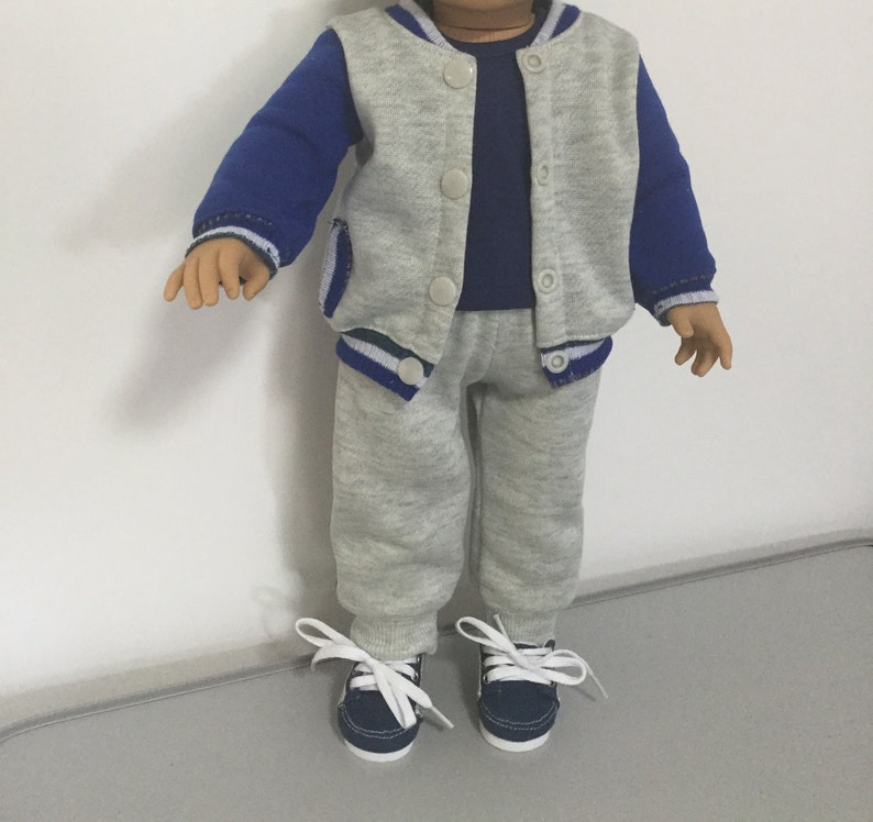May include: A gray and blue tracksuit for a doll. The jacket has a blue and white stripe on the sleeves and a white stripe on the bottom. The pants are gray with a white stripe on the bottom. The doll is wearing navy blue sneakers.