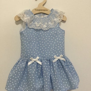 Puede incluir: Un vestido azul claro con estampado de corazones blancos y ribete de encaje blanco. El vestido tiene lazos blancos en la parte delantera y está colgado en una percha de madera con el texto "Valspiersssews".