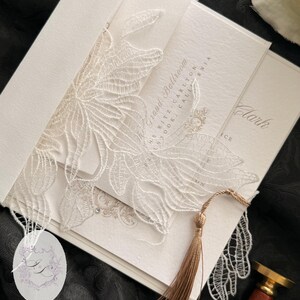 Puede incluir: Conjunto de invitaciones de boda en marfil con intrincados detalles de encaje floral. La invitaci&oacute;n incluye una borla, y el texto "The Grand Ballroom" y "Clark" es visible. El conjunto se presenta sobre una superficie negra.