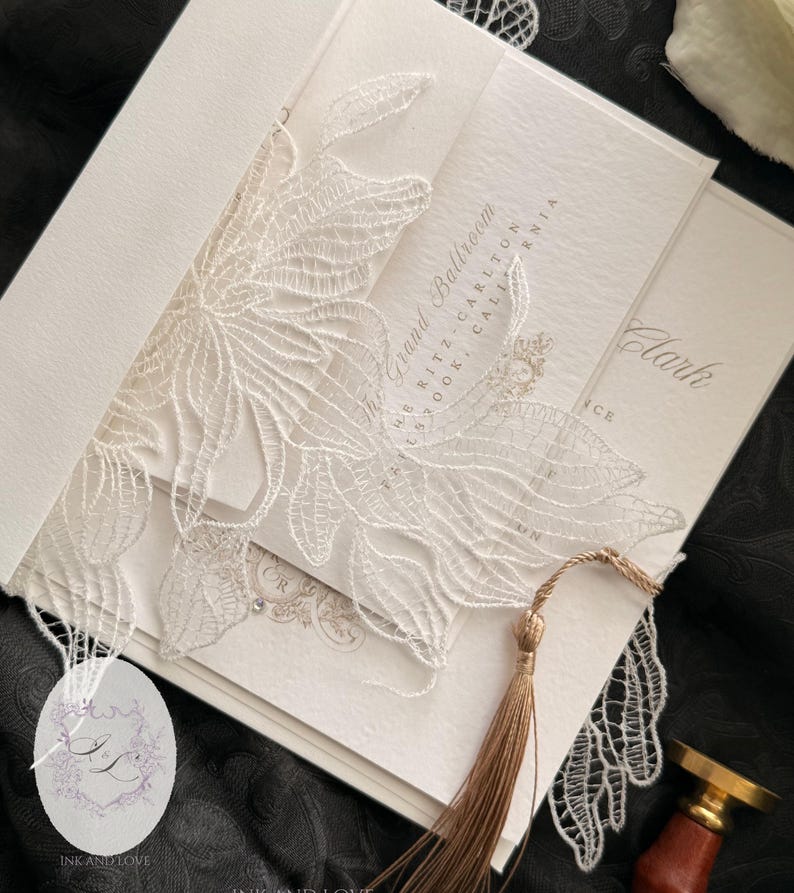 Puede incluir: Conjunto de invitaciones de boda en marfil con intrincados detalles de encaje floral. La invitaci&oacute;n incluye una borla, y el texto "The Grand Ballroom" y "Clark" es visible. El conjunto se presenta sobre una superficie negra.