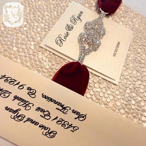Może przedstawiać: Eleganckie zaproszenie ślubne z imionami "Rose & Ryan" w ozdobnym kroju pisma. Zaproszenie zawiera kremową kartę z błyszczącą ozdobą z cyrkonii i wstążką z ciemnoczerwonego aksamitu. Na karcie wydrukowana jest data "02/25/2026".