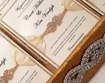 May include: Elegant wedding invitations with gold glitter accents and cream-colored ribbons. Each invitation features a decorative jeweled embellishment. The text includes names and a date, suggesting a formal event.