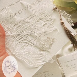 Puede incluir: Elegante invitaci&oacute;n de boda con intrincados detalles de encaje blanco. La invitaci&oacute;n presenta los nombres "Mitte & Ryan Clark" e incluye una borla decorativa. El dise&ntilde;o es cl&aacute;sico y sofisticado, perfecto para un evento formal.