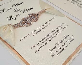 May include: Ivory wedding invitation with a rose gold border and a jeweled embellishment. The invitation includes the names "Rose White & Ryan Clark" and the event details. A matching envelope is visible in the background.