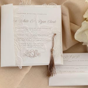 Puede incluir: Elegante invitaci&oacute;n de boda blanca con delicados detalles de encaje y una borla. La invitaci&oacute;n presenta los nombres "White & Ryan Clark" e informaci&oacute;n de RSVP. Un dise&ntilde;o sofisticado para una ocasi&oacute;n especial.