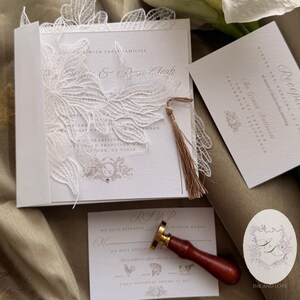 Puede incluir: Elegante juego de invitaciones de boda blanco con intrincados detalles de encaje. Incluye invitaci&oacute;n, tarjeta RSVP, tarjeta de recepci&oacute;n y un sello de lacre. La invitaci&oacute;n presenta los nombres "Kate & Ryan Lark" y detalles del evento.