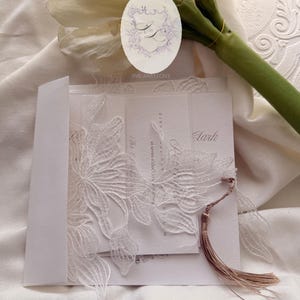 Puede incluir: Elegante invitaci&oacute;n de boda blanca con intrincados detalles de encaje floral. La invitaci&oacute;n presenta una borla y va acompa&ntilde;ada de un sobre a juego. El dise&ntilde;o incluye el texto "Ink and Love" y otros detalles del evento.