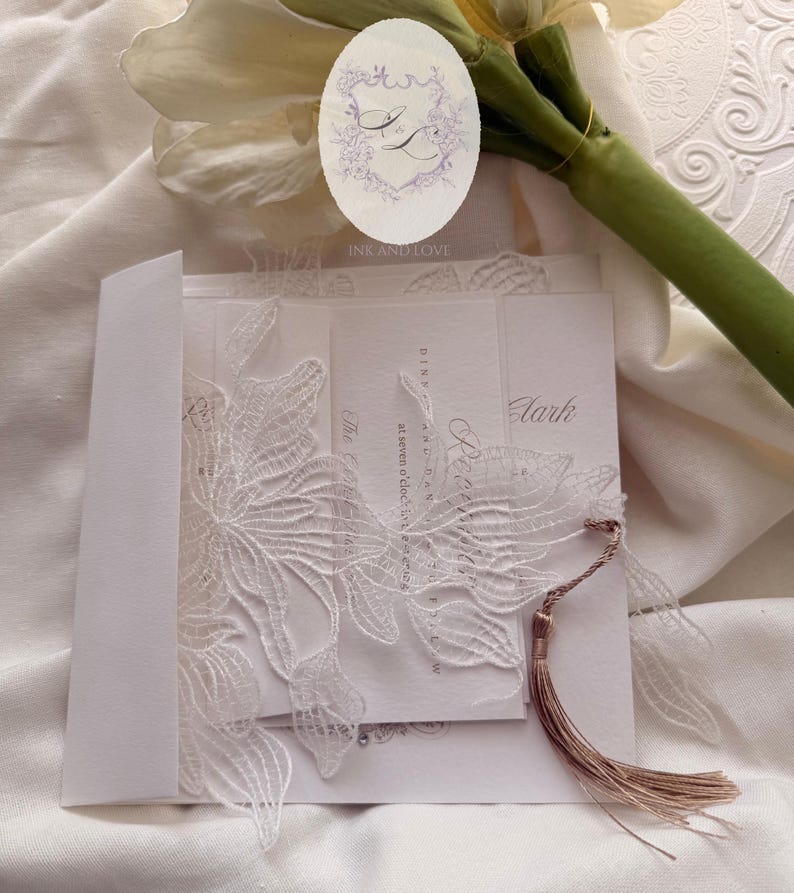 Puede incluir: Elegante invitaci&oacute;n de boda blanca con intrincados detalles de encaje floral. La invitaci&oacute;n presenta una borla y va acompa&ntilde;ada de un sobre a juego. El dise&ntilde;o incluye el texto "Ink and Love" y otros detalles del evento.