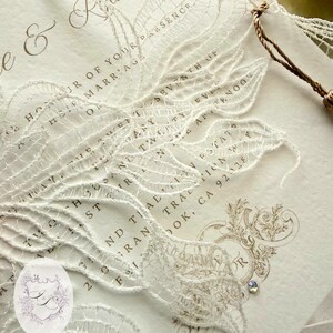 Puede incluir: Invitaci&oacute;n de boda marfil con delicada superposici&oacute;n de encaje blanco. La invitaci&oacute;n presenta una escritura elegante y elementos decorativos, incluyendo una peque&ntilde;a joya. El texto incluye los nombres "e & Ryan" y la fecha.