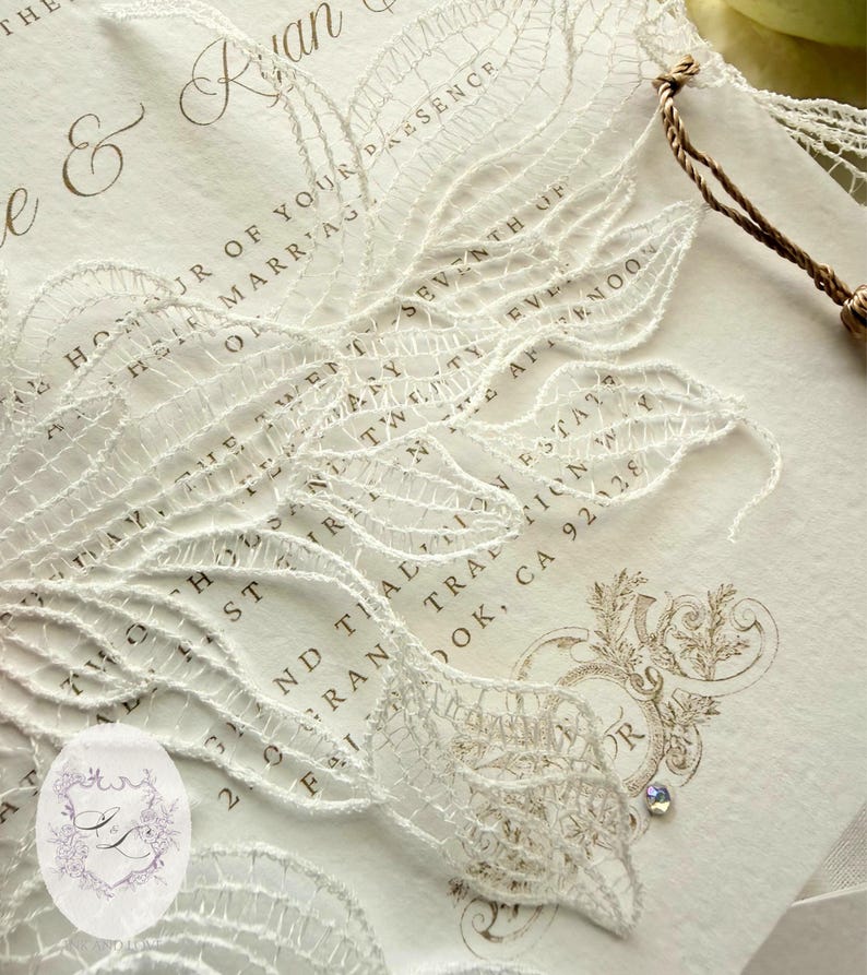 Puede incluir: Invitaci&oacute;n de boda marfil con delicada superposici&oacute;n de encaje blanco. La invitaci&oacute;n presenta una escritura elegante y elementos decorativos, incluyendo una peque&ntilde;a joya. El texto incluye los nombres "e & Ryan" y la fecha.