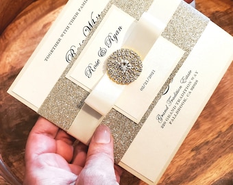 May include: An ivory wedding invitation with gold glitter accents and a cream-colored ribbon. The invitation features the names "Rose & Ryan" and the date "02/21/2021". A decorative jewel embellishes the ribbon.