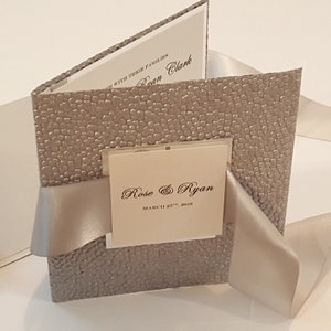 Invitación de boda de lujo con bolsillo desplegable, en relieve blanco y plateado, con lazo de satén llamativo, monograma y tarjeta RSVP. Fabricada en EE. UU. Envíos a todo el mundo.