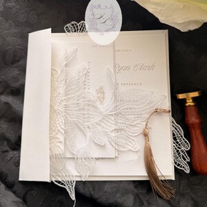 Puede incluir: Invitaci&oacute;n de boda marfil con una superposici&oacute;n de encaje blanco. La invitaci&oacute;n presenta el nombre "Ryan Clark" y est&aacute; acentuada con una borla dorada. Un sello de cera y un sobre completan el elegante dise&ntilde;o.