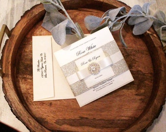 May include: A wedding invitation with the names "Rose White" and "Rose & Ryan" is displayed on a wooden tray. The invitation features a white ribbon and a sparkling embellishment. An envelope and decorative greenery are also present.