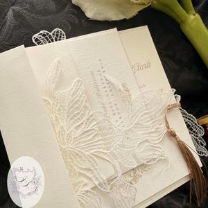 Puede incluir: Conjunto de invitaciones de boda marfil con una delicada superposici&oacute;n de encaje blanco. La invitaci&oacute;n presenta texto dorado y una borla. El texto de la invitaci&oacute;n dice "Sky Grand Ballroom" y "Clark". El logotipo "INK AND LOVE" tambi&eacute;n es visible.
