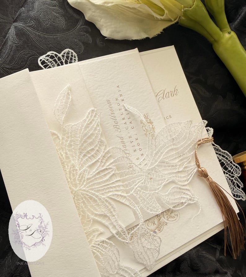 Puede incluir: Conjunto de invitaciones de boda marfil con una delicada superposici&oacute;n de encaje blanco. La invitaci&oacute;n presenta texto dorado y una borla. El texto de la invitaci&oacute;n dice "Sky Grand Ballroom" y "Clark". El logotipo "INK AND LOVE" tambi&eacute;n es visible.