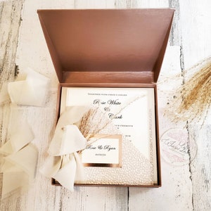 May include: A wedding invitation suite in a brown box. The invitation features the names "Rose White & Clark" and "Rose & Ryan" with the date "03/25/21". The suite includes a cream-colored invitation, a decorative bow, and dried floral accents.