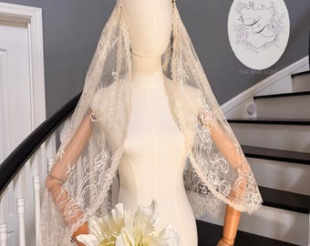 Puede incluir: Un velo de encaje de color crema drapeado sobre la cabeza y los hombros de un maniquí. El velo presenta un patrón floral con bordes festoneados. Un ramo de flores blancas se coloca en la base del maniquí. Un logotipo con el texto INK AND LOVE.