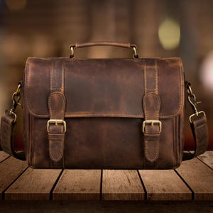 May include: A dark brown leather satchel featuring a top handle, a fold-over flap with two vertical straps, and brass-toned metal buckles. The bag includes detachable side shoulder straps with swivel clips and visible stitching along the edges.