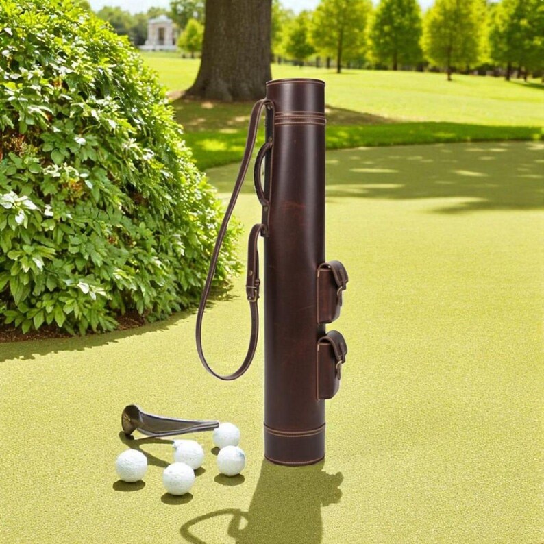 May include: A brown leather golf club carrier with a shoulder strap and two small pouches. Several white golf balls and a golf club rest on the green surface. The carrier is cylindrical and stands upright.
