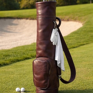 May include: A brown leather golf bag with golf clubs, a white towel, and golf gloves. Two golf balls and tees are on the green grass. The bag has a shoulder strap and a side pocket.