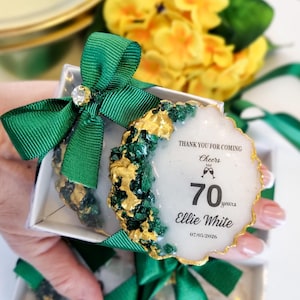 Puede incluir: Un conjunto de recuerdos de fiesta en cajas transparentes, cada uno atado con una cinta verde y un pedrería. Los recuerdos son blancos con detalles dorados y verdes, y presentan el texto "Thank you for coming", "Cheers", "70 years", "Ellie White", y la fecha.