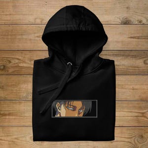 May include: Black hooded sweatshirt with an embroidered anime character's eyes and part of the face. The design is in a rectangular frame. The hoodie is folded on a wooden surface.