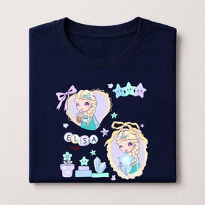 May include: Navy blue t-shirt with a cartoon illustration of Elsa from Frozen. The design includes Elsa with a drink, stars, and the name "ELSA" in colourful letters. Additional elements include a flower pot and crystals.