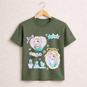 May include: Olive green t-shirt with a cartoon illustration of Elsa from Frozen. The design includes Elsa in two heart-shaped frames, stars, and the name "ELSA" in letter blocks. The shirt has a crew neck and short sleeves.