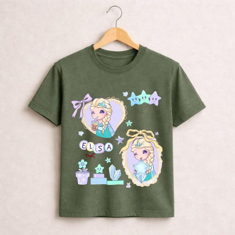 May include: Olive green t-shirt with a cartoon illustration of Elsa from Frozen. The design includes Elsa in two heart-shaped frames, stars, and the name "ELSA" in letter blocks. The shirt has a crew neck and short sleeves.