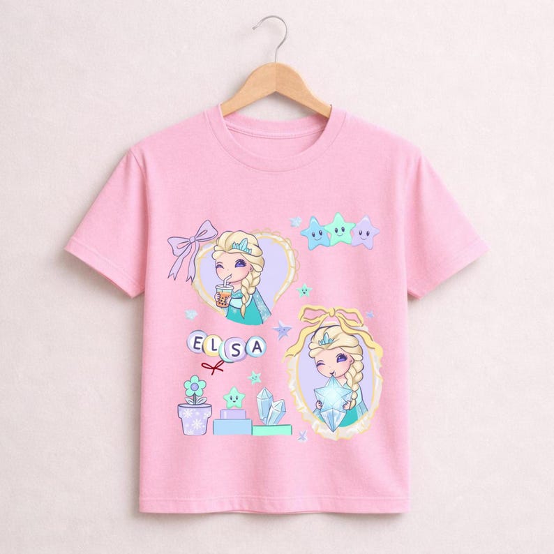 May include: A pink t-shirt with a cartoon illustration of Elsa from Frozen. The design includes Elsa's portrait, the word "ELSA", stars, and other decorative elements. The shirt is suspended from a wooden hanger.