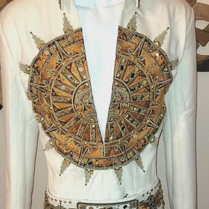 May include: White jumpsuit with elaborate gold and bronze embellishments. The jacket features a large, circular design on the chest, and the belt has a decorative buckle. The outfit is displayed on a mannequin.