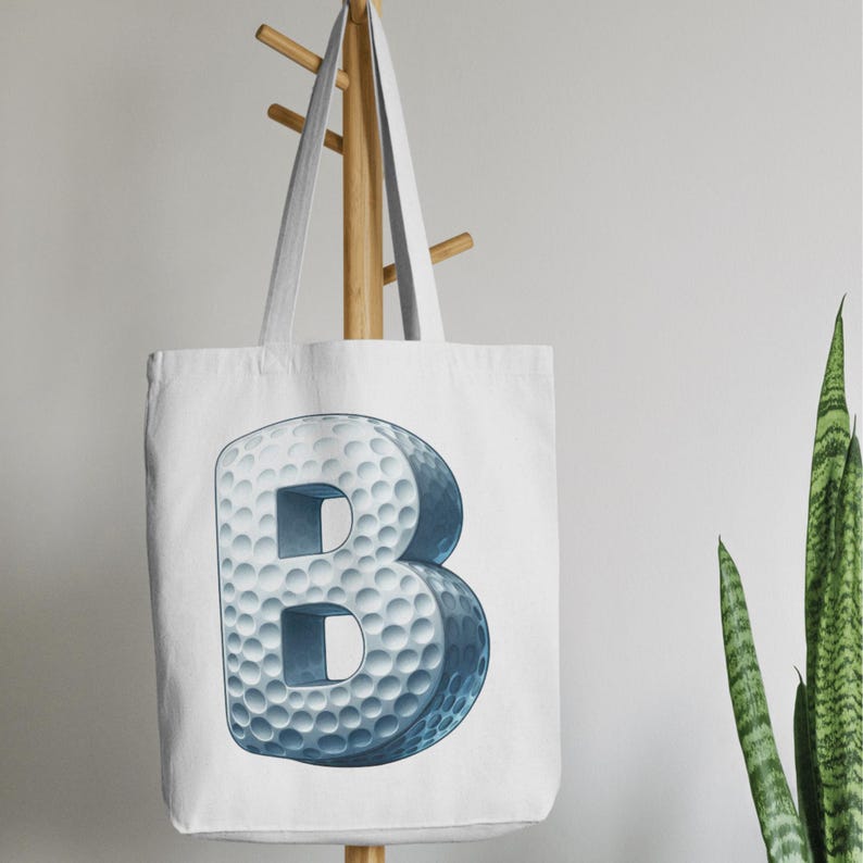 Op de afbeelding: Witte draagtas met een grote, 3D-letter "B" die is ontworpen om op een golfbal te lijken. De letter is lichtblauw met een putjesstructuur en de tas heeft lichtgrijze bandjes. De tas hangt aan een houten rek.