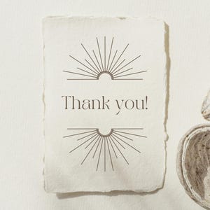 Op de afbeelding: Een cr&egrave;mekleurige kaart met een geschulpte rand met de woorden "Thank you!" in een schreefletter. Boven en onder de tekst staan symmetrische sunburst-ontwerpen in donkerbruin. De kaart heeft een minimalistisch, elegant ontwerp.