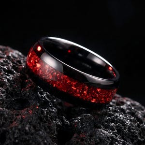 Puede incluir: Un anillo negro con una banda pulida y una incrustación central de material rojo triturado que tiene un aspecto brillante y texturizado. El anillo está colocado sobre una superficie oscura, porosa y rocosa con una textura negra rugosa.