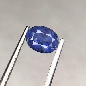May include: A faceted, oval-shaped, deep blue gemstone. The gem is held by silver tweezers, showcasing its polished surface and internal facets. The stone's color ranges from a rich, dark blue to lighter shades, creating a play of light.