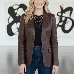 May include: A brown leather blazer with a classic design. The blazer is paired with a black top and dark blue jeans. A gold necklace adds a touch of elegance. The background features abstract art.