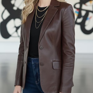 May include: A brown leather blazer with a notched lapel and two-button closure. The blazer has a tailored fit and a single chest pocket. Worn with a black top, dark blue jeans, and a gold necklace.