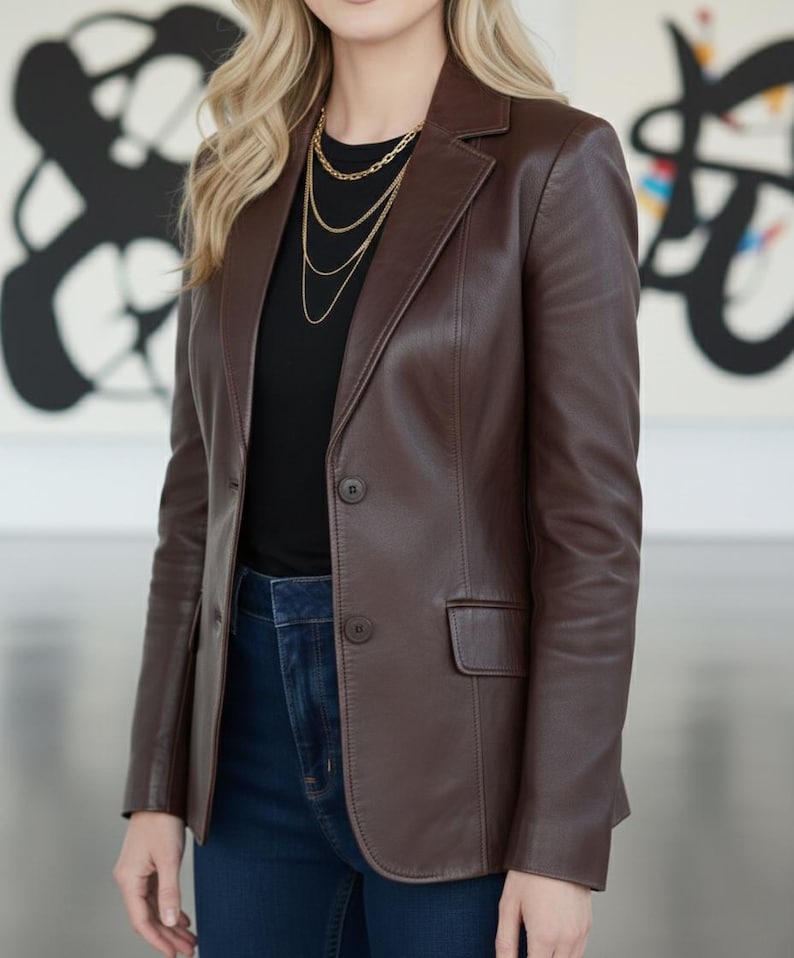May include: A brown leather blazer with a notched lapel and two-button closure. The blazer has a tailored fit and a single chest pocket. Worn with a black top, dark blue jeans, and a gold necklace.