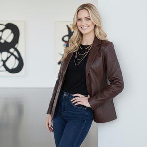 May include: A brown leather blazer paired with a black top and dark blue jeans. The blazer has a classic design. A gold necklace adds a touch of elegance. The outfit is suitable for various occasions.