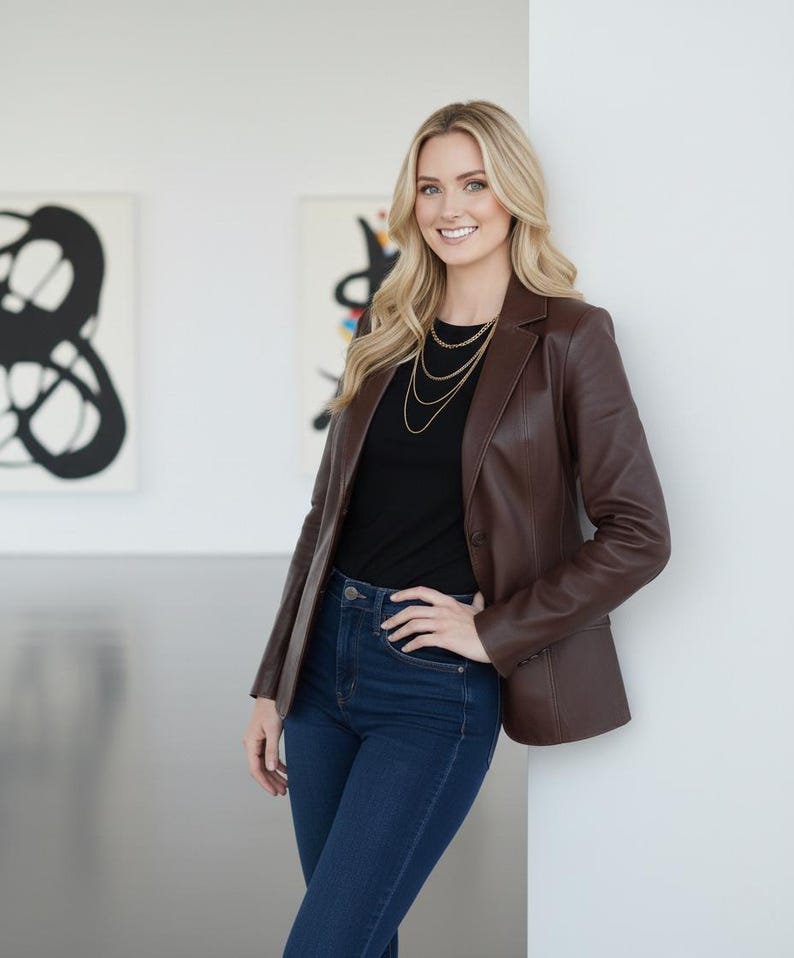 May include: A brown leather blazer paired with a black top and dark blue jeans. The blazer has a classic design. A gold necklace adds a touch of elegance. The outfit is suitable for various occasions.