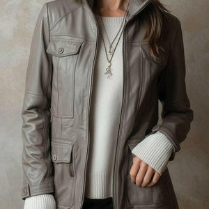 Puede incluir: Chaqueta de cuero gris con cremallera, múltiples bolsillos y cuello alzado. La chaqueta se lleva sobre un suéter blanco. Las mangas del suéter son visibles en las muñecas. Se lleva un collar plateado.