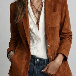 May include: A tan suede blazer with two front pockets and gold buttons. The blazer is worn over a white button-down shirt and blue jeans. The model is also wearing layered necklaces and bracelets.