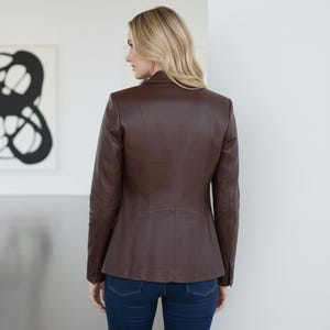 May include: A brown leather blazer with a tailored fit, featuring a notched collar and buttoned cuffs. The jacket is paired with dark blue denim jeans. The model is facing away from the camera.