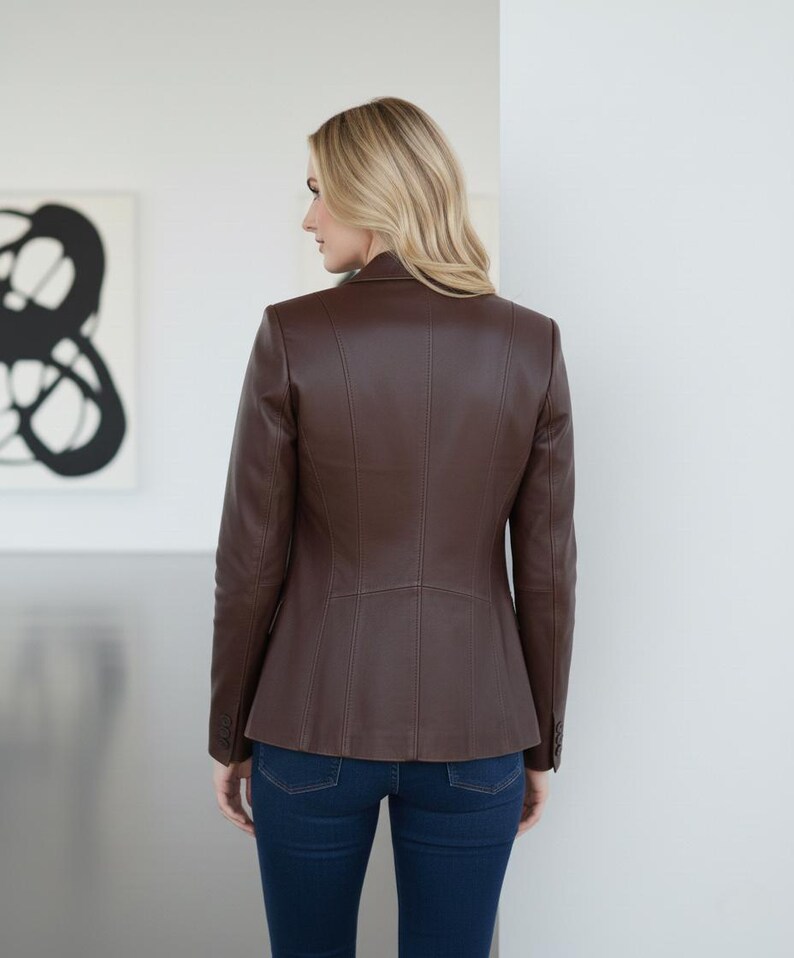 May include: A brown leather blazer with a tailored fit, featuring a notched collar and buttoned cuffs. The jacket is paired with dark blue denim jeans. The model is facing away from the camera.